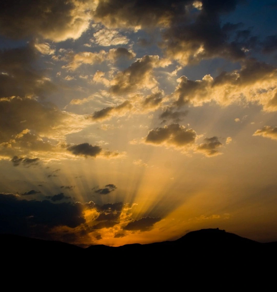 Sunset with golden rays piercing through dramatic clouds over silhouetted mountains.