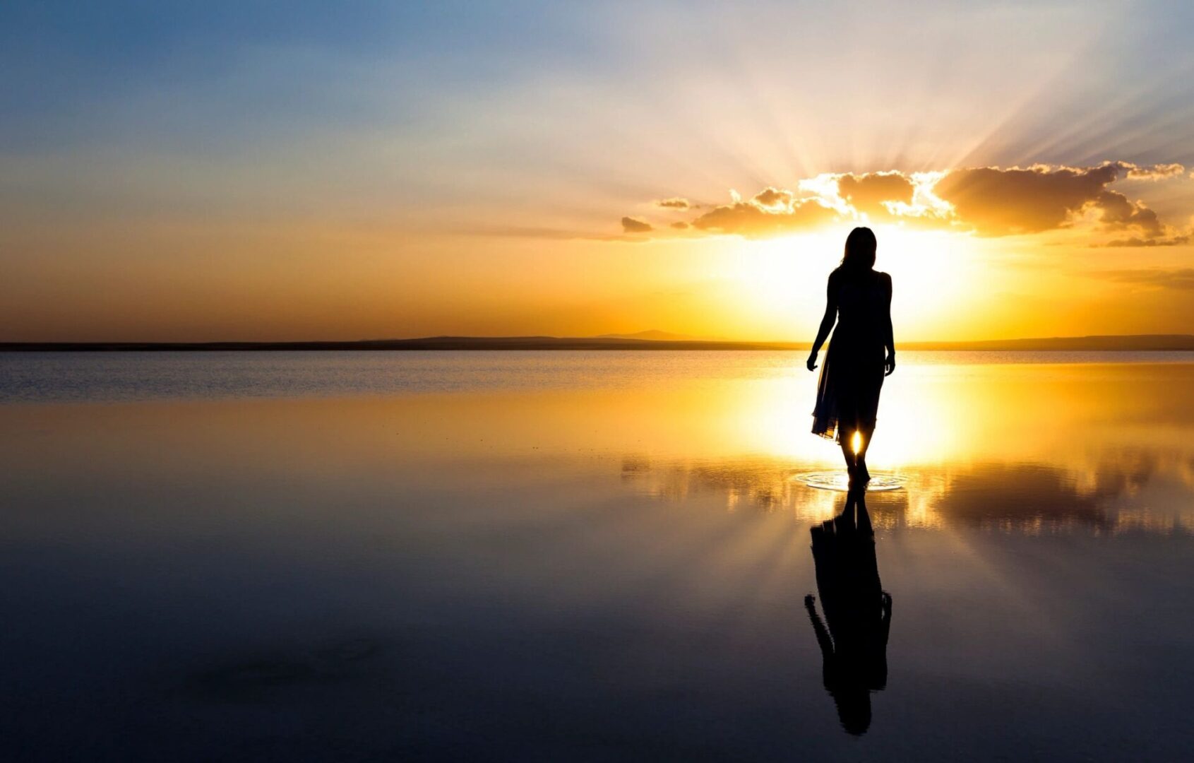 A person walking along a serene beach at sunset, casting a reflection on the wet sand.